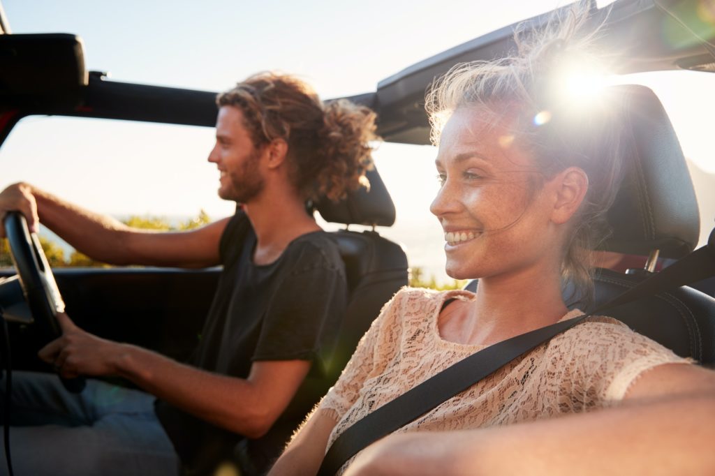 Millennial white couple on a road trip driving in an open top car, backlit, close up